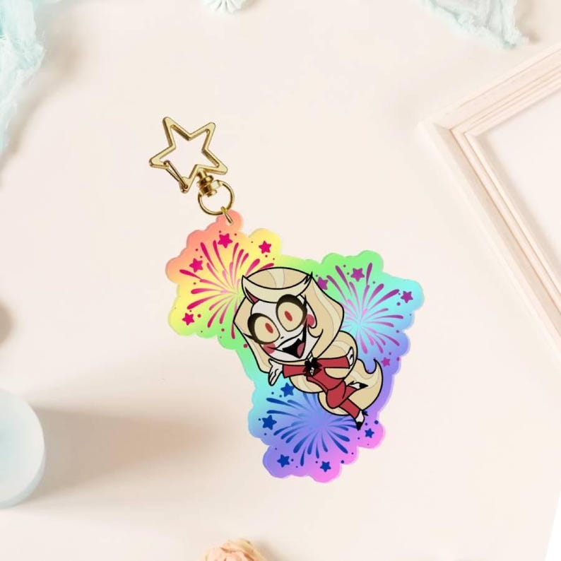 May include: An acrylic keyring featuring a cartoon character with blonde hair and a red outfit, set against a vibrant rainbow-coloured background with firework and star patterns. The charm is attached to a gold-coloured star-shaped metal clasp.