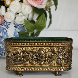 May include: An oval brass planter featuring a raised relief pattern of blooming roses and leaves. The container has a beaded border along the top and bottom edges, a dark green interior lining, and stands on four small, detailed claw-style feet.