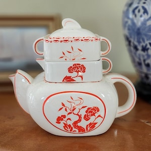May include: A white ceramic teapot set with red floral designs. The set includes a teapot, a lid, and two stackable rectangular cups. The teapot has a rounded shape with a curved handle and spout. The floral design is repeated on the cups and lid.