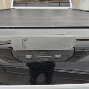 May include: A black truck bed cover with a gray, textured, rectangular panel. The panel has a small, black footprint logo and two black rectangular buttons.