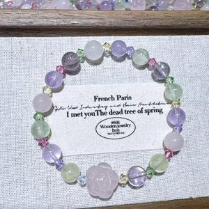 May include: A bracelet with pastel-coloured beads, including lavender, pink, and green, and a rose-shaped charm. The bracelet has crystal accents and the text "French Paris" and "I met you The dead tree of spring" is visible. The bracelet is a delicate piece.