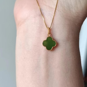 May include: A gold-colored necklace featuring a four-leaf clover pendant with a green center. The clover is outlined in gold and hangs from a delicate gold chain. The necklace is displayed against a skin-toned background.
