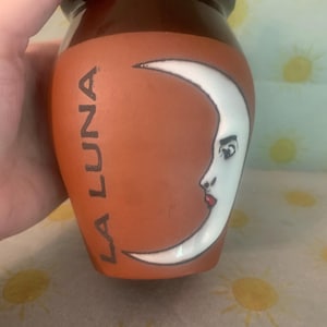 May include: A terracotta-colored ceramic vase with a dark brown glazed rim. The body features a white crescent moon graphic with a human face, including a visible eye and red lips. The text LA LUNA is printed vertically in black lettering.