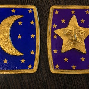 May include: Two rectangular plaques with gold trim. One features a gold crescent moon on a blue background with gold stars. The other has a gold star with a face on a purple background, also with gold stars. The plaques are likely decorative.