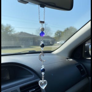 May include: A car charm featuring a silver chain with a decorative arrangement of beads and charms. The charm includes a clear heart-shaped crystal, a crescent moon, and various blue and silver beads. The charm hangs from the rearview mirror.