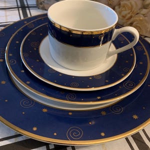 May include: A white porcelain teacup and saucer set stacked on a matching dinner plate. The pieces feature a deep navy blue border decorated with gold-colored spiral and star patterns, finished with a thin gold rim along the edges of each item.