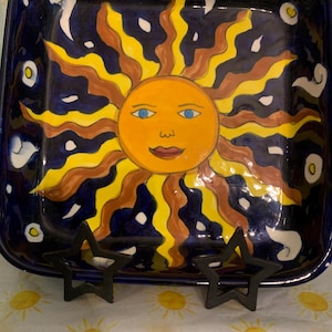 May include: A square ceramic dish featuring a hand-painted yellow sun with a face, blue eyes, and wavy orange and yellow rays. The background is a deep navy blue decorated with small white cloud-like shapes and yellow circles, supported by two black star-shaped stands.