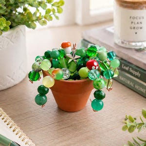 May include: A small terracotta pot filled with a beaded plant arrangement. The arrangement features various shades of green beads, including round and faceted shapes, with a few red and orange accents. A silver star charm hangs from the arrangement.