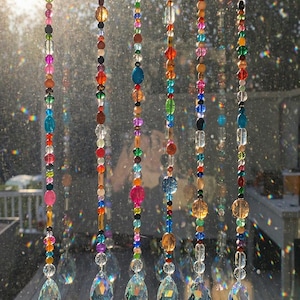 May include: Handmade glass crystal beaded suncatchers in various colors, including pink, blue, orange, and clear. Each strand features a teardrop-shaped crystal at the bottom. The text reads "Throws Rainbows in Sun".