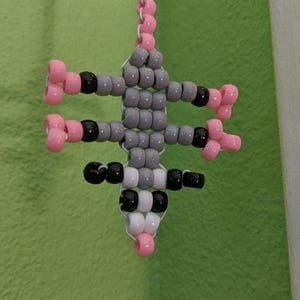 May include: A gray, black, and pink beaded charm in the shape of a rat. The charm is hanging from a white beaded chain.
