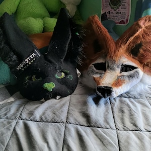 May include: Two animal masks, one black with Monster Energy branding and green accents, and one orange, white, and black fox-like mask. Both masks feature pointed ears, mesh eye coverings, and detailed fur-like textures on their surfaces.