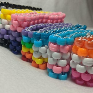 Könnte beinhalten: Ein Set aus bunten Perlenarmbändern, jedes mit einem anderen Muster. Die Armbänder sind aus kleinen, runden Perlen in verschiedenen Farben gefertigt, darunter Schwarz, Grau, Weiß, Rosa, Lila, Blau, Grün, Gelb und Orange. Die Armbänder sind übereinander gestapelt und bilden so eine optisch ansprechende Darstellung.