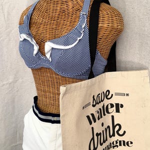 May include: A blue and white gingham bikini top with white ruffle trim, white shorts with a navy and white striped waistband, and a beige canvas tote bag featuring the black text "save water drink champagne Austin, TX 2019".