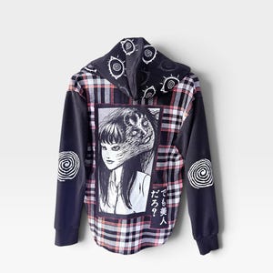 May include: A black and white plaid hoodie featuring a graphic print of a woman with a distorted face on the back. The hood and sleeves are decorated with white eye and spiral patterns. The Japanese text reads, "でも美人だろ？"