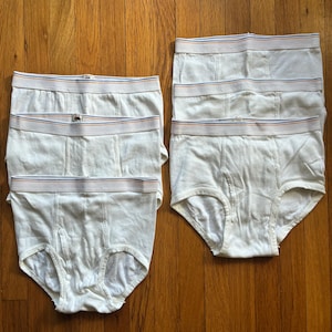 May include: A set of six white briefs arranged in two columns. Each pair of underwear features a thick elastic waistband with thin horizontal blue and orange stripes. The briefs are constructed with a standard fly front design and leg openings.