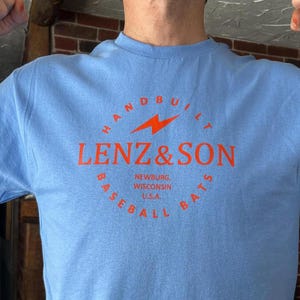 May include: A light blue t-shirt with a red circle logo that says "HANDBUILT LENZ & SON BASEBALL BATS" with a lightning bolt graphic and the text "NEWBURG, WISCONSIN U.S.A."
