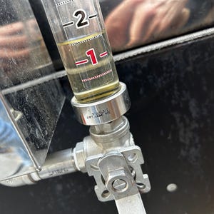 May include: A close-up of a silver valve with a clear glass tube attached. The tube has a red line and markings for "1" and "2". The tube is filled with a yellow liquid.
