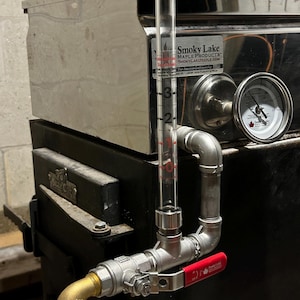 May include: A stainless steel maple syrup evaporator with a clear glass sight gauge, a red valve handle, and a gauge with a black face and a white needle. The gauge reads "Smoky Lake Maple Products" and "smokylakmaple.com".