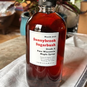 May include: A clear glass bottle filled with amber-colored maple syrup. The label reads "Sunnybrook Sugarbush, Grade A Pure Wisconsin Maple Syrup." The bottle is sealed with a black cap and contains 16 fl oz (473 ml).
