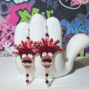 May include: A pair of novelty earrings featuring a cartoon character with red hair and a surprised expression. The earrings have silver hooks and dangling red and pink beads. The background is a white hand-shaped display against a graffiti wall.
