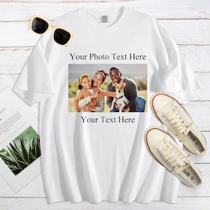 Pode incluir: Camiseta branca com foto de fam&iacute;lia e sobreposi&ccedil;&atilde;o de texto. A foto mostra uma fam&iacute;lia com um cachorro. &Oacute;culos de sol e t&ecirc;nis de lona est&atilde;o ao lado da camiseta. Uma revista tamb&eacute;m est&aacute; na imagem.