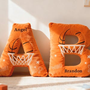 May include: Two orange plush letter cushions, "A" and "B", with basketball designs. Each cushion has a basketball, hoop, and net. The letter "A" has the name "Angel", and "B" has "Brandon". Ideal for a child's bedroom.