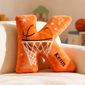 Puede incluir: Cojín naranja de felpa con forma de letra "K" con diseño de baloncesto. El cojín presenta una pelota, una red y el nombre "Kevin". Tiene detalles de estrellas y trofeos, ideal para una habitación deportiva.