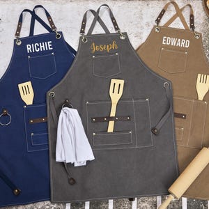 Puede incluir: Tres delantales de lona personalizados con correas ajustables, cada uno con un nombre diferente: Richie, Joseph y Edward. Cada delantal tiene un bolsillo grande con una espátula de madera y un bolsillo más pequeño con un anillo de metal. Los delantales son de color azul, gris y marrón.
