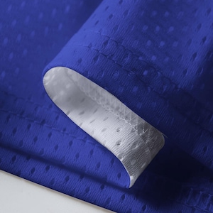 May include: Close-up of a royal blue fabric with a mesh-like texture. The edge of the fabric is folded over, revealing a white interior. The fabric has small, evenly spaced holes, creating a breathable material.