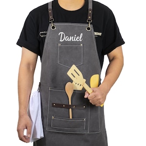 Puede incluir: Un delantal de lona gris con correas de cuero marrón y un bolsillo grande. El delantal tiene un nombre, "Daniel", bordado en la parte delantera. El delantal lo lleva una persona que sostiene una espátula de madera y una cuchara de madera.