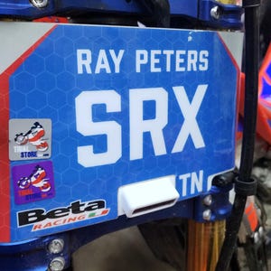 May include: A blue and white motorcycle number plate with the text "RAY PETERS SRX TN" and a Beta Racing logo. The plate is attached to a red and blue motorcycle.
