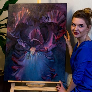 Könnte beinhalten: Originales Gemälde auf Leinwand, das eine Nahaufnahme einer großen Irisblüte zeigt. Das Kunstwerk zeigt Farbtöne von Lila, Rosa und Blau mit einem dunklen Hintergrund. Das Gemälde wird auf einer Holzstaffelei präsentiert.