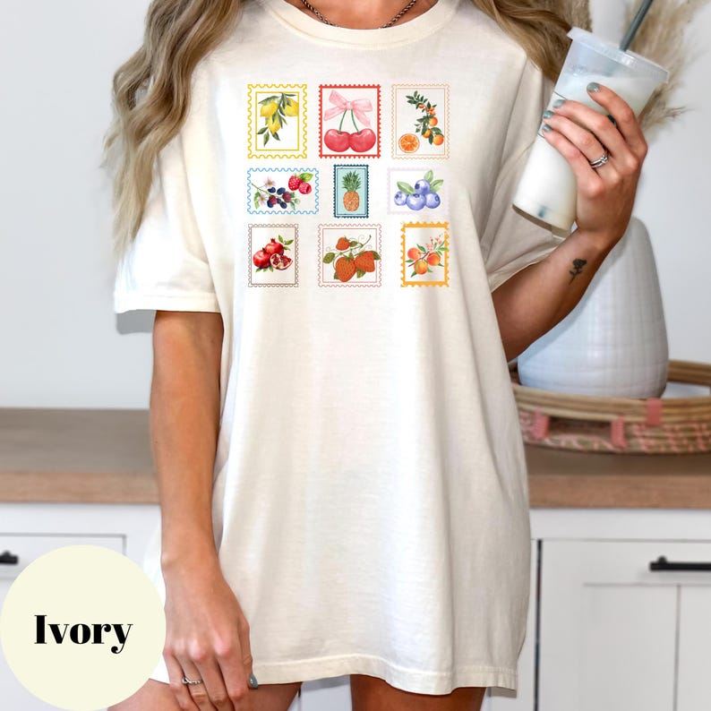 May include: Ivory-colored t-shirt featuring a collection of colorful fruit postage stamp designs. The stamps showcase illustrations of lemons, cherries, oranges, berries, pineapple, blueberries, pomegranates, strawberries, and flowers. The shirt is a relaxed fit.