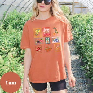 May include: A rust-colored t-shirt featuring a grid of fruit-themed postage stamp designs. The stamps showcase various fruits like lemons, cherries, and blueberries. The shirt is a short-sleeved, crew neck style.