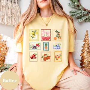 May include: Butter-colored t-shirt featuring a collection of fruit postage stamp designs. The stamps showcase illustrations of lemons, cherries, oranges, berries, pineapple, blueberries, pomegranates, strawberries, and peaches. The shirt has a relaxed fit.