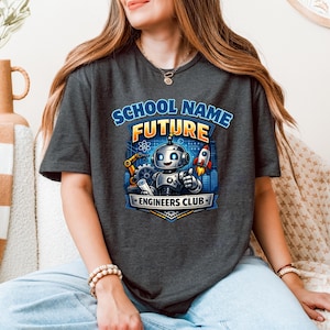 Peut inclure: T-shirt gris foncé avec un graphisme coloré représentant un robot, une fusée et des engrenages. Le texte sur le t-shirt indique "SCHOOL NAME FUTURE ENGINEERS CLUB". Le design est dans les tons de bleu, orange et argent.