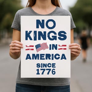 Op de afbeelding: Een wit bord met de woorden "NO KINGS IN AMERICA SINCE 1776" in blauwe tekst, samen met een Amerikaanse vlag. Het bord wordt vastgehouden door een persoon die een grijs t-shirt draagt.