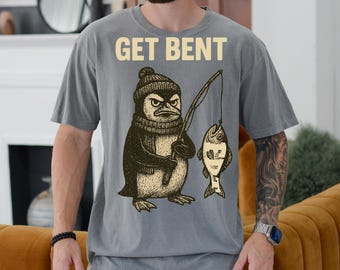 Puede incluir: Una camiseta gris con un gráfico de un pingüino con un sombrero y sosteniendo una caña de pescar con un pez en la línea. El texto "GET BENT" está impreso en la camiseta.