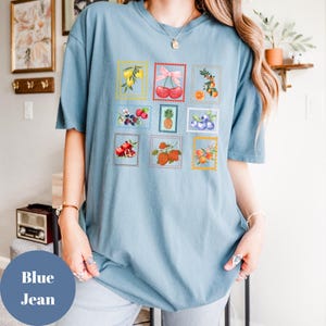 May include: Blue jean t-shirt featuring a collection of fruit illustrations in postage stamp designs. The stamps showcase various fruits like lemons, cherries, oranges, strawberries, and blueberries. The shirt has a relaxed fit and short sleeves.