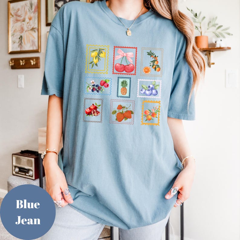 May include: Blue jean t-shirt featuring a collection of fruit illustrations in postage stamp designs. The stamps showcase various fruits like lemons, cherries, oranges, strawberries, and blueberries. The shirt has a relaxed fit and short sleeves.