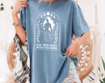 May include: A light blue t-shirt with a white graphic of a woman with long hair standing in a crescent moon arch. The text "She who rises with the moon" is printed below the graphic.