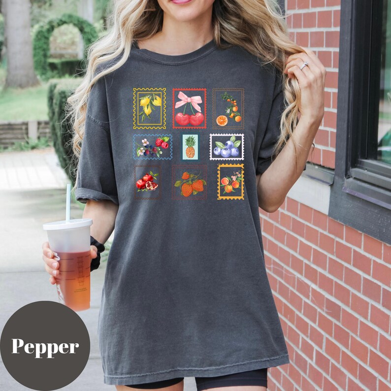 May include: Dark gray t-shirt featuring a collection of colorful fruit illustrations within postage stamp frames. The stamps showcase various fruits like cherries, oranges, and blueberries. The word "Pepper" is printed at the bottom of the image.