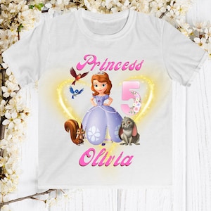 May include: White t-shirt with a pink princess design featuring a cartoon princess, a squirrel, a rabbit, and a bird. The text "Princess" and "Olivia" are in pink, and the number "5" is in pink with flowers.