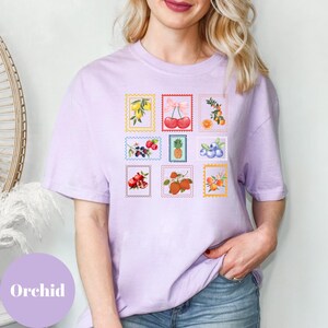 May include: A light purple t-shirt featuring nine colorful fruit-themed postage stamp designs. The stamps showcase illustrations of lemons, cherries, oranges, berries, a pineapple, blueberries, pomegranates, strawberries, and apricots. The word "Orchid" is printed on the bottom left.