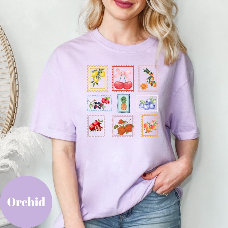 May include: A light purple t-shirt featuring nine colorful fruit-themed postage stamp designs. The stamps showcase illustrations of lemons, cherries, oranges, berries, a pineapple, blueberries, pomegranates, strawberries, and apricots. The word "Orchid" is printed on the bottom left.