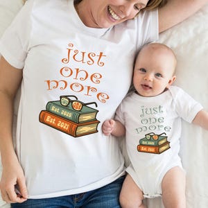 Puede incluir: Una camiseta blanca con el texto "just one more" y una pila de libros con gafas en la parte superior. El texto "Est. 2025" y "Est. 2021" está en los libros. Un bebé lleva un body blanco con el mismo texto y diseño de libro.