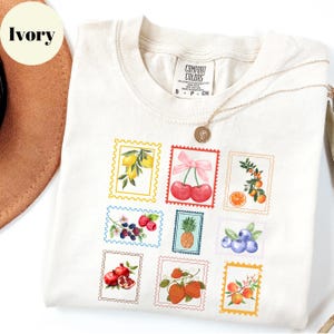 May include: Ivory-colored t-shirt with a crew neck, featuring a collection of colorful fruit postage stamp designs. The stamps include lemons, cherries, raspberries, a pineapple, blueberries, pomegranates, and oranges. A gold necklace with a pendant is draped over the shirt.
