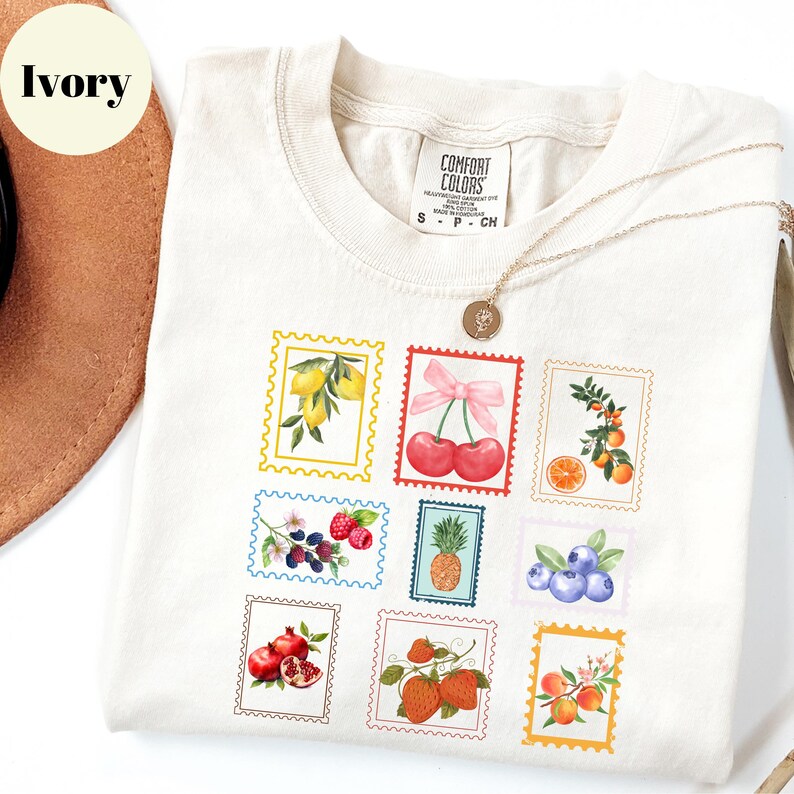 May include: Ivory-colored t-shirt with a crew neck, featuring a collection of colorful fruit postage stamp designs. The stamps include lemons, cherries, raspberries, a pineapple, blueberries, pomegranates, and oranges. A gold necklace with a pendant is draped over the shirt.