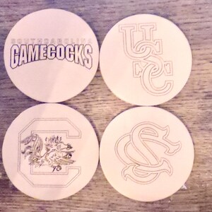 May include: Four round coasters with university logos and text. One coaster reads "SOUTH CAROLINA GAMECOCKS" in purple and white. Other coasters feature the letters "USC" and "SC" in a stylized design, and a Gamecock logo.