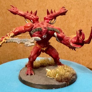 May include: A red, four-armed demon miniature with a sword and a base covered in brown and green grass.